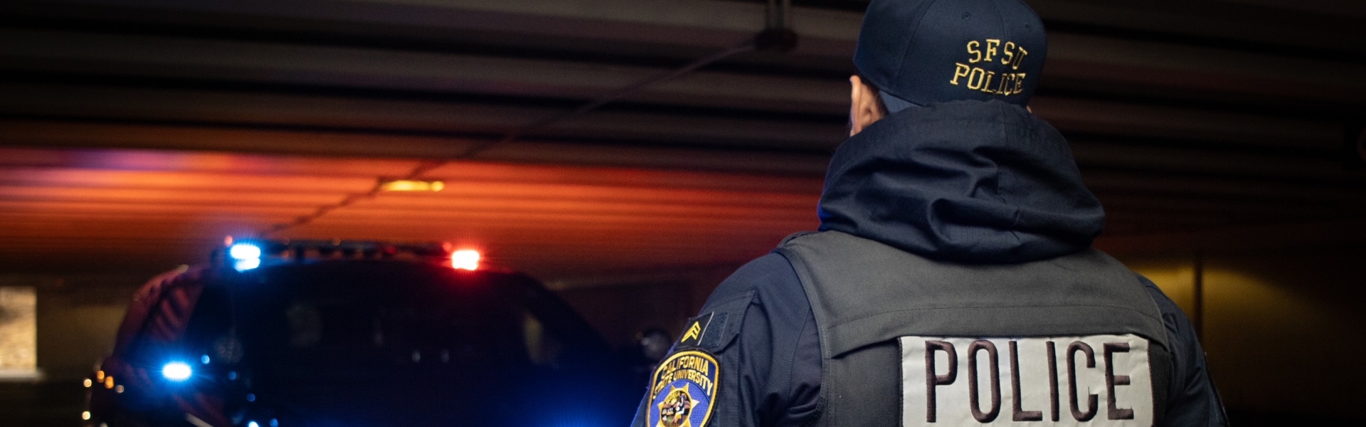 Image of the back of a UPD officer standing in front of UPD Police SUV with lights